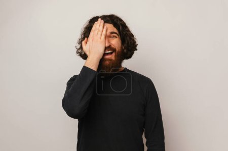 Photo for Bearded man with long hair hold a palm on a half of his face while smiling over grey background. - Royalty Free Image