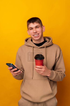 Foto de Retrato de un joven feliz hombre con capucha sosteniendo teléfono inteligente y taza de café para llevar. - Imagen libre de derechos