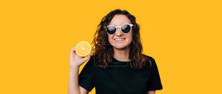 Téléchargez les photos : Portrait studio d'une femme tenant la moitié d'une orange sur fond jaune disant que l'été est là bébé. - en image libre de droit