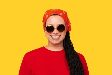 Studioaufnahme eines breit lächelnden Mädchens mit Zöpfen aus Schachteln mit Sonnenbrille vor gelbem Hintergrund.