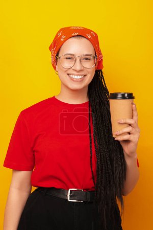 Tiro vertical de una chica sonriente con trenzas de caja sosteniendo una taza de café para llevar.