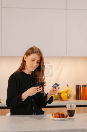 Mujer joven concentrada está comprando en línea durante el uso de teléfono y tarjeta de crédito. Ella se queda en su cocina en casa.