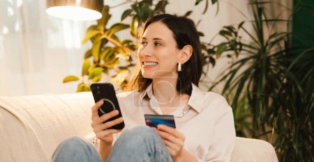 A woman is happily using her smartphone and credit card to shop online while relaxing on a cozy sofa at home
