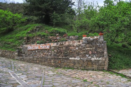 The village name 'Barcena Mayor' displayed on a rustic stone wall at the village entrance, with cobblestone pavement leading into the green landscape