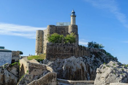 Téléchargez les photos : Le phare historique de Castro Urdiales, qui fait partie d'un ancien château, se dresse sur une falaise escarpée sur fond de ciel dégagé - en image libre de droit