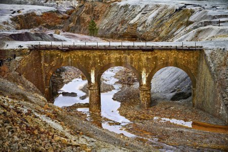 An old arch bridge crosses over a stream tinged with the reddish hues of pollution in the Rio Tinto mining area