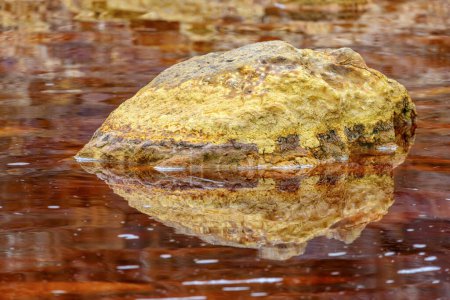 A solitary golden rock casts its reflection upon the tranquil, iron-rich red waters of Rio Tinto