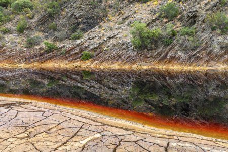 Markante Erdschichten und ein kräftiger Streifen rotes Wasser säumen den rissigen Boden des Rio Tinto