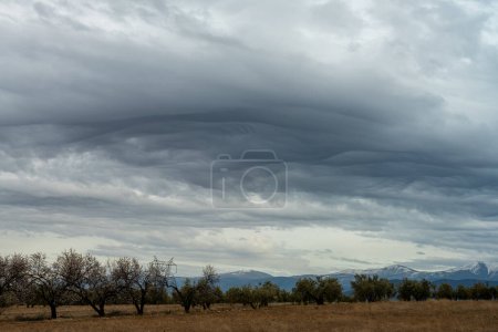 Dramatic asperitas clouds blanket the sky above a serene rural landscape with distant mountains, creating a striking natural scene