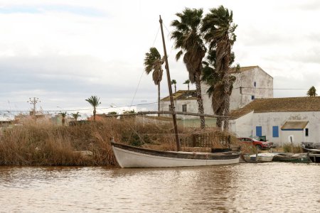A traditional sailing boat rests by the reeds on the banks of Albufera Valencia, with rustic buildings and palm trees in the background