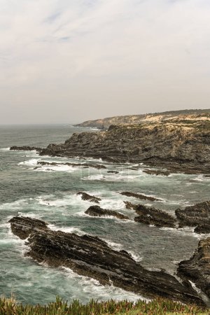 Majestic cliffs at Cabo Sardao, Portugal, with rugged rock formations and waves crashing against the shoreline. A partly cloudy sky enhances the dramatic coastal scenery.
