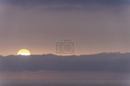 Ein heiterer Sonnenaufgang über dem Ozeanhorizont, wobei die Sonne teilweise von einer niedrigen Wolkenbank verdeckt wird. Die sanften Pastellfarben des Himmels schaffen eine friedliche und ruhige Szenerie.