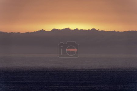 A serene sunrise over the ocean horizon, with the sun partially hidden by a low cloud bank. The soft pastel colors of the sky create a peaceful and tranquil scene.