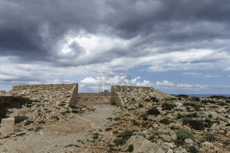 Une batterie antiaérienne abandonnée à Cabo de Cavalleria avec des nuages sombres et dramatiques au-dessus. Les ruines historiques se dressent dans un paysage accidenté.