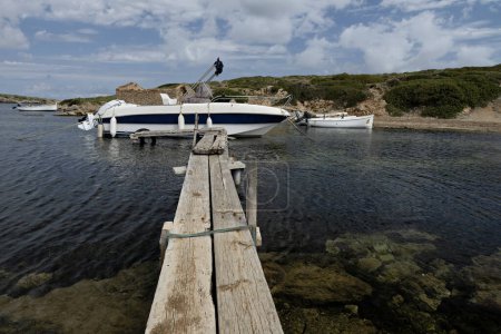 Wooden pier leading to boats docked in the clear waters of Port of Sanitja, Menorca. Scenic view of the rocky shoreline and lush greenery under a partly cloudy sky.