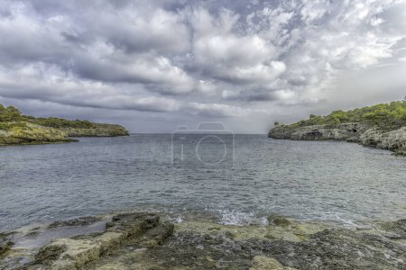 Picturesque scene of Cala Turqueta in Menorca, showcasing clear waters, rocky shoreline, and lush greenery. The tranquil bay is framed by a partly cloudy sky.