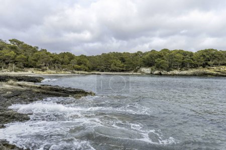 Scène pittoresque de Cala Turqueta à Minorque, présentant des eaux claires, un littoral rocheux et une végétation luxuriante. La baie tranquille est encadrée par un ciel partiellement nuageux.