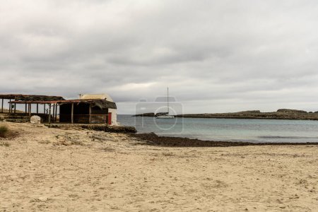 A rustic beach hut stands by the rocky shoreline of Cala Binibeca, Menorca, under a cloudy sky.