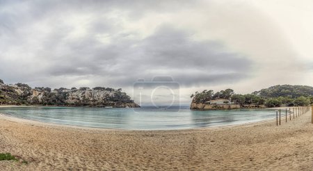 Una vista serena de la playa de Cala Galdana en Menorca, con aguas tranquilas y acantilados costeros. El primer plano de la playa de arena conduce a una pintoresca cala enmarcada por promontorios rocosos y exuberante vegetación.