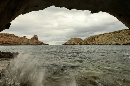 Dramatic view from inside a coastal cave at Cala Morell, Menorca, showcasing rugged rocky formations and crashing waves under a cloudy sky.