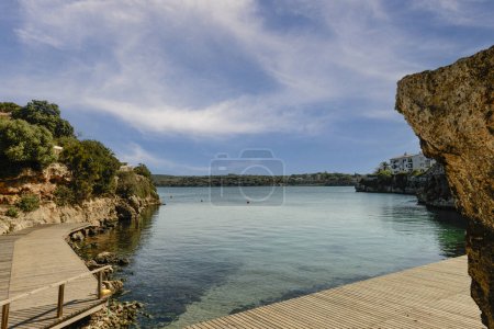 A serene view of Playa Secreta cove in Menorca, featuring clear blue waters, rocky cliffs, and a wooden pathway leading to the water's edge under a bright blue sky.