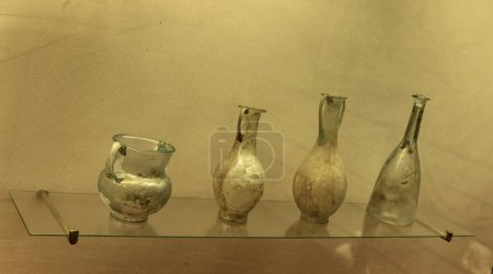 Un antiguo jarrón de cristal verde romano con un diseño intrincado. Cuenta con un cuerpo ancho, un cuello estrecho y asas decorativas, que muestran la artesanía de la fabricación de vidrio romano.