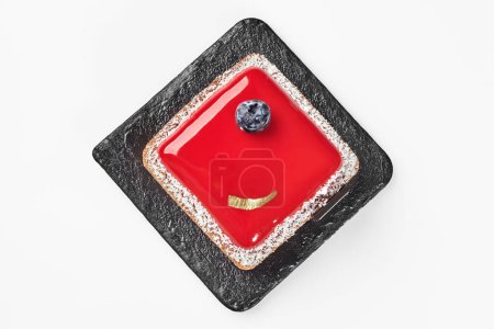Téléchargez les photos : Vue du dessus du petit gâteau au fromage carré garni d'une couche de gelée de baies, saupoudré de flocons de noix de coco garni de bleuets mûrs et d'or comestible servi sur une planche d'ardoise noire sur fond blanc - en image libre de droit