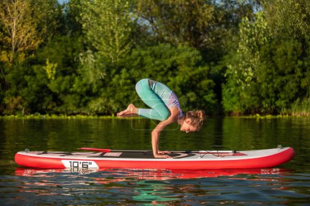 Téléchargez les photos : Jeune femme font du yoga sur un stand up paddle board SUP sur un beau lac ou une rivière. Le concept d'un mode de vie sain. Du sport. Du yoga. hobby - en image libre de droit