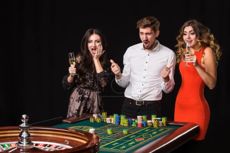 Dos mujeres jóvenes y un hombre detrás de la mesa de ruleta sobre fondo negro. Emociones jugadores