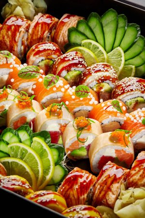 Foto de Primer plano de varios apetitosos rollos de sushi japoneses con jengibre en escabeche, pepino y rodajas de limón, verduras envasadas en caja de cartón negro. Auténtica cocina. Entrega o comida para llevar concepto - Imagen libre de derechos