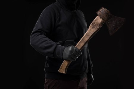 Foto de Un hombre con guantes sostiene un hacha en sus manos contra un fondo negro. Criminal. - Imagen libre de derechos