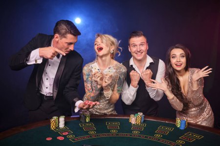 Téléchargez les photos : Group of a happy rich buddies are playing poker at casino. Youth are making bets waiting for a big win. They are looking happy standing at the table against a red and blue backlights on black - en image libre de droit