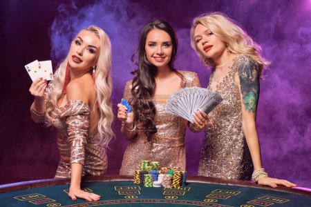 Charming females with a perfect hairstyles and bright make-up, dressed in a golden shiny dresses are posing standing at a gambling table. Poker concept on a black smoke background with pink and blue