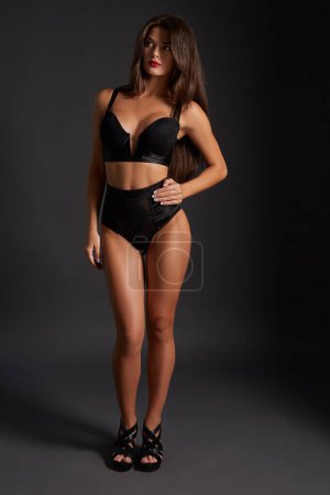Photo for Young sexy slim tanned woman in black underwear posing against dark grey background. Fashion portrait of beautiful girl with long wavy brunette hair. Underwear or bikini model - Royalty Free Image