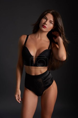 Téléchargez les photos : Stunning caucasian female model with dark hair and red lips in black underwear posing on dark grey background. Fashion portrait of young pretty beautiful brunette woman. - en image libre de droit