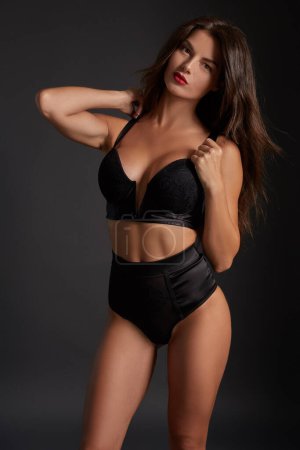 Photo for Stunning caucasian female model with dark hair and red lips in black underwear posing on dark grey background. Fashion portrait of young pretty beautiful brunette woman. - Royalty Free Image