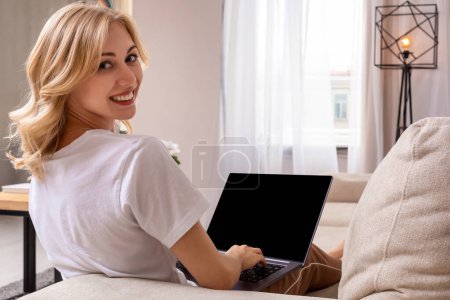 Photo for Woman on sofa working on laptop with mockup blank screen. Empty copy space on monitor for advertisement, language learning ad, online shopping website, social media site. Black screen. - Royalty Free Image