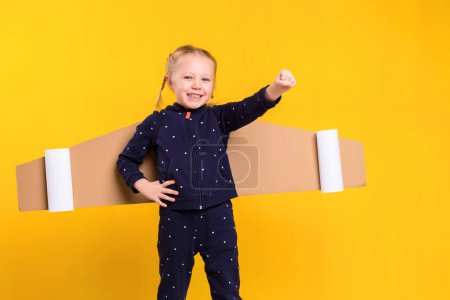 Photo for A little child girl is wearing homemade cardboard flying wings, pretending to be a pilot for a craft, imagination or exploration concept. Studio photography on a yellow background - Royalty Free Image