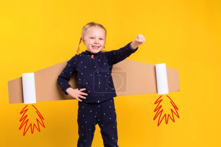 Photo for A little child girl is wearing homemade cardboard flying wings, pretending to be a pilot for a craft, imagination or exploration concept. Studio photography on a yellow background - Royalty Free Image