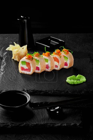 Photo for Delicious rolls filled with tuna, salmon, avocado and cream cheese wrapped in mamenori topped with spicy sauce and tobiko served on black slate board with wasabi, gari and soy sauce. Japanese snack - Royalty Free Image