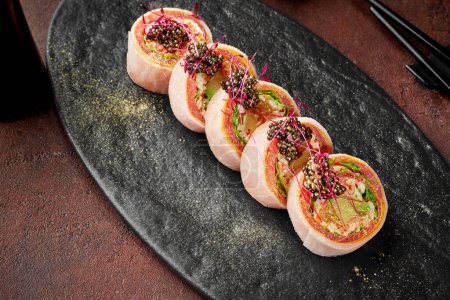 Foto de Apetitivos rollos de sushi sin arroz con atún, tortilla japonesa, lechuga, aguacate y naranja envueltos en mamenori adornados con tobiko negro espolvoreado con polvo de oro servido en pizarra - Imagen libre de derechos