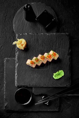 Foto de Conjunto de rollos de sushi envueltos en mamenori rematados con tobiko y virutas de cebolletas servidas tradicionalmente con wasabi, gari y salsa de soja en pizarra, vista superior sobre fondo negro. Comida japonesa - Imagen libre de derechos