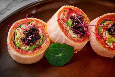 Foto de Rollos de sushi con carne de cangrejo, aguacate, lechuga y rodaja de naranja envueltos en atún, tortilla japonesa y mamenori sin arroz adornado con tobiko negro y microverde servido en plato. Cocina japonesa - Imagen libre de derechos