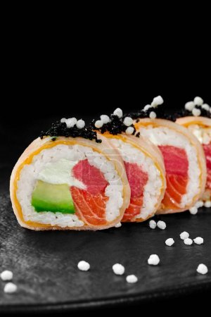 Foto de Primer plano de delicados rollos de sushi envueltos en mamenori rellenos de atún crudo, salmón, aguacate y queso crema rematados con tobiko negro y bolas de arroz crujiente servido en pizarra. Comida japonesa - Imagen libre de derechos