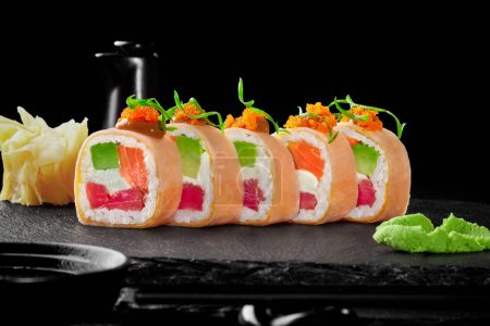 Photo for Gourmet sushi rolls with raw tuna, salmon, avocado and cream cheese wrapped in mamenori with spicy sauce and tobiko caviar served on black slate board with wasabi and gari. Japanese seafood delicacies - Royalty Free Image