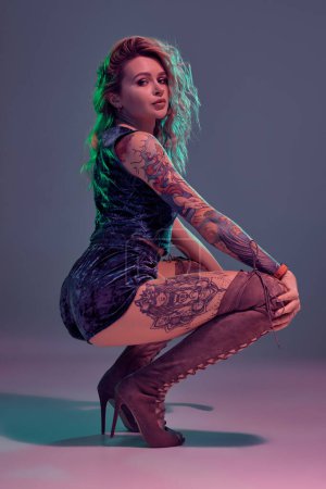 Photo for Young beautiful tattooed blonde woman wearing blue velour booty shorts and t-short, maroon suede boots, showing some twerk moves, looks seductive. - Royalty Free Image