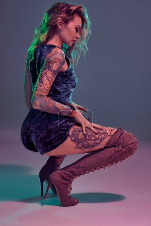 Young beautiful tattooed blonde woman wearing blue velour booty shorts and t-short, maroon suede boots, showing some twerk moves, looks seductive.
