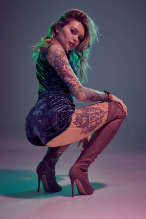 Photo for Young beautiful tattooed blonde woman wearing blue velour booty shorts and t-short, maroon suede boots, showing some twerk moves, looks seductive. - Royalty Free Image