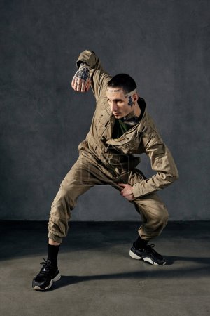 Young modern fellow with tattooed body and face, earrings, beard. Dressed in khaki jumpsuit and black sneakers. He dancing against gray studio background. Dancehall, hip-hop. Full length, copy space
