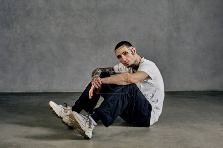 Athletic fellow with tattooed body and face, beard. Dressed in white t-shirt and sneakers, black sports trousers. Sitting sideways on floor against gray background. Dancehall, hip-hop. Close up